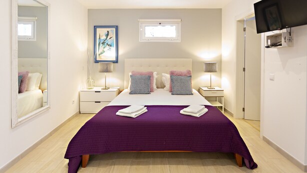 A modern bedroom with a large bed, two bedside tables, a flat-screen TV, and a framed artwork on the wall. - Talabriga - Apt Cidade (Tavira)