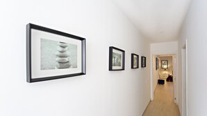 A hallway with framed artwork on the walls, leading to a room with a bed and a nightstand. - Talabriga - Apt Cidade (Tavira)