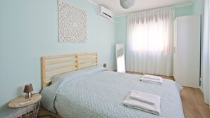 Un dormitorio con cama de madera, una mesita de noche con lámpara, aire acondicionado de pared y un amplio ventanal con cortinas.