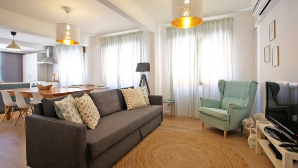 Una sala de estar moderna con un sofá gris, un sillón azul verdoso y un televisor sobre un mueble blanco. Hay un comedor con una mesa y sillas, y una cocina al fondo.
