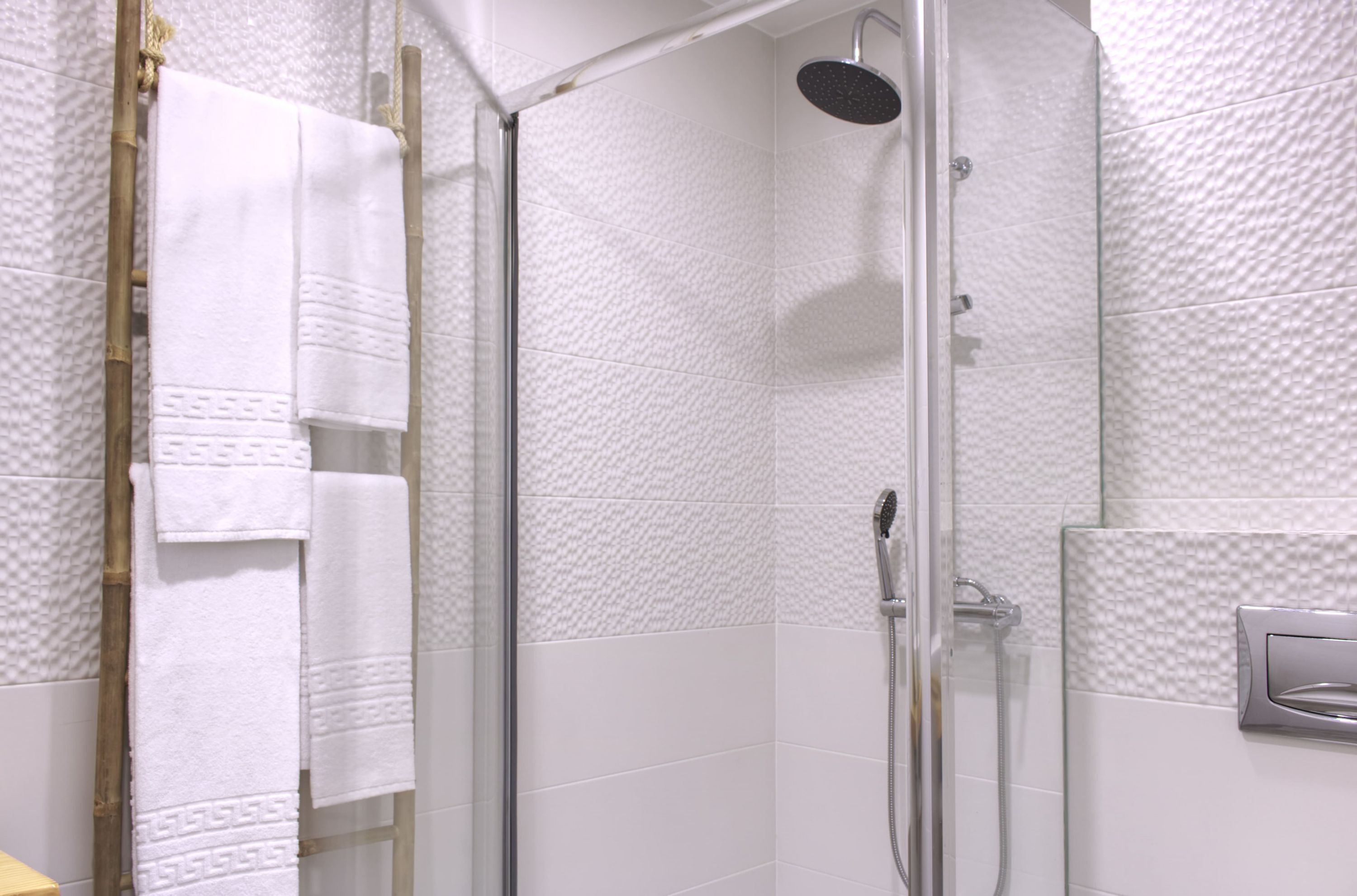 A glass-enclosed shower with a handheld showerhead, white textured walls, and a wooden ladder.