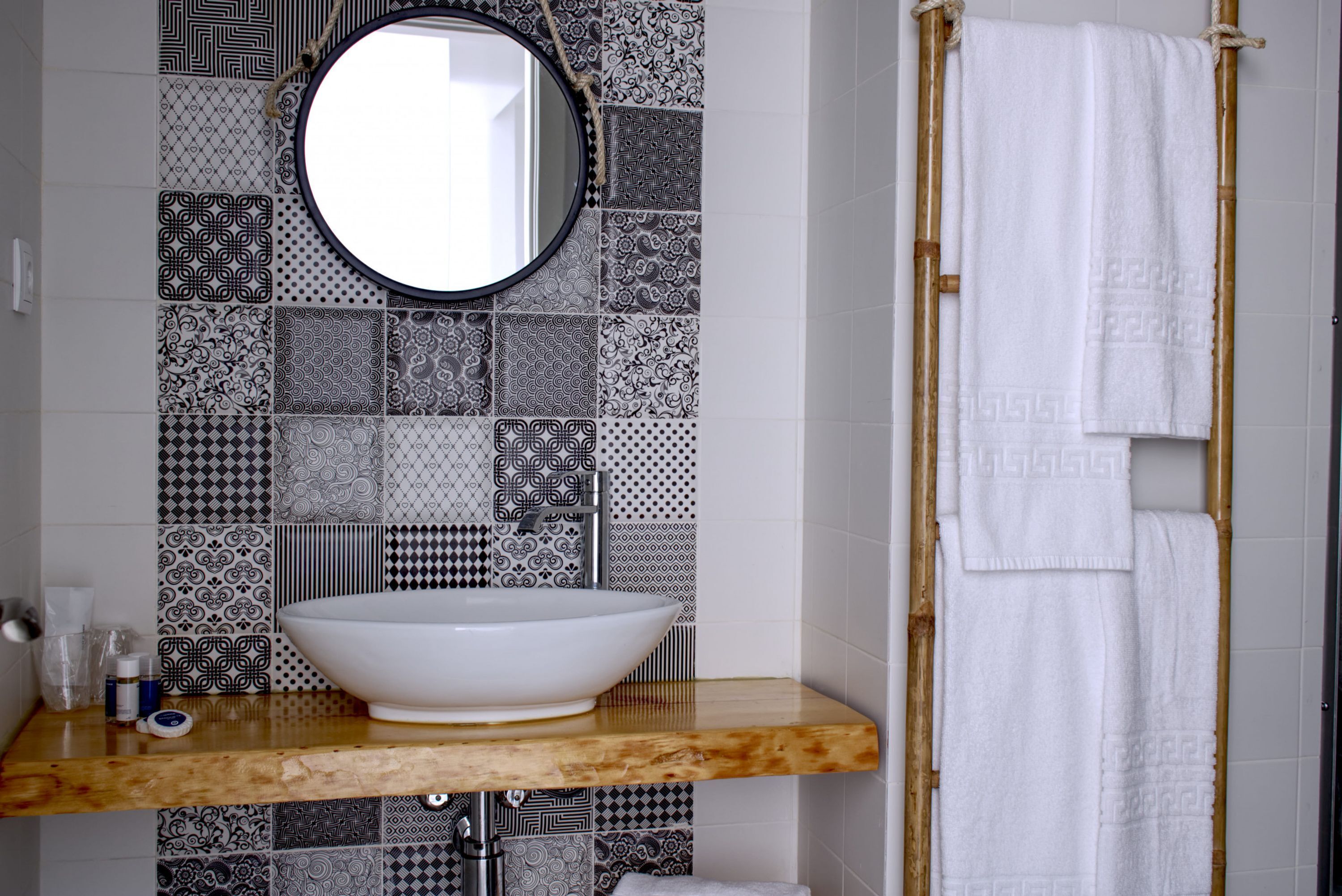 A bathroom with a white vessel sink, a round mirror, and a towel rack with two white towels. The wall is decorated with black and white patterned tiles.