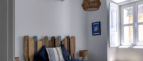 A bedroom with a wooden headboard, a blue bedspread, a wicker hanging light, a framed picture on the wall, and a window with daylight.