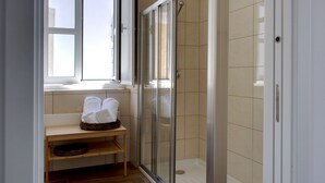 A bathroom with a glass shower enclosure, a wooden bench with towels, and a window providing natural light.