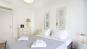 A neatly made bed with folded white towels, a bedside table with a lamp, and decorative wall art. - Cabanas Terrace in Tavira (Tavira)