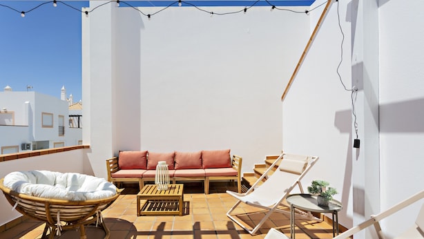 A sunlit balcony with a white sofa, a wicker chair, a wooden table, and a small potted plant. - Cabanas Terrace in Tavira (Tavira)