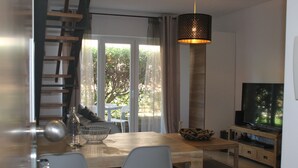 Een moderne woonkamer met een houten eettafel, twee stoelen, een televisie op een houten dressoir en een groot raam met uitzicht op bomen.
