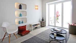 A modern living room with a white chair, a small white table, a bookshelf, and a large window with a view of the outdoors. - Casa Dona Ana in Tavira (Tavira)
