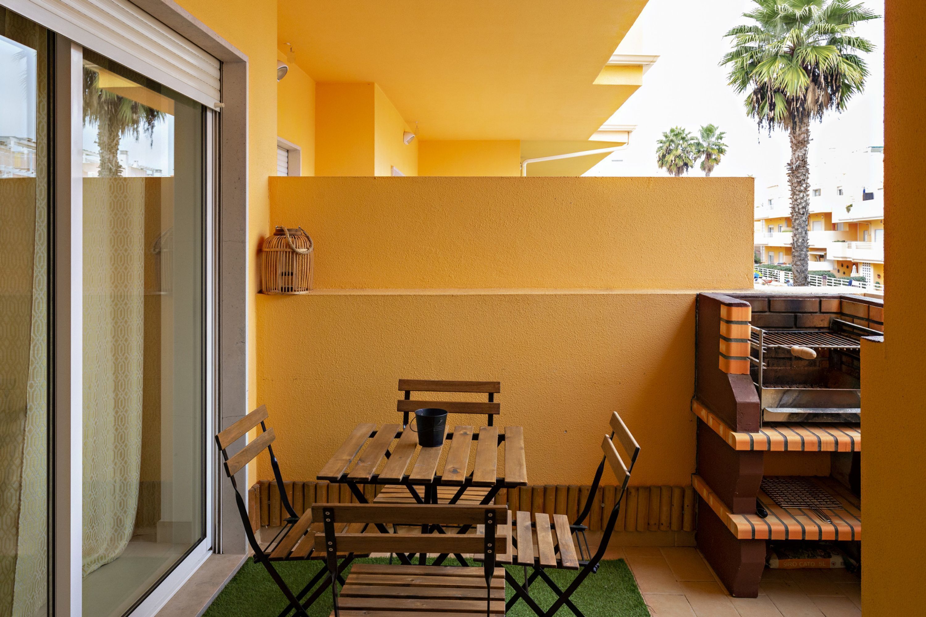 A balcony with a wooden table and chairs, a grill, and a view of palm trees and buildings.