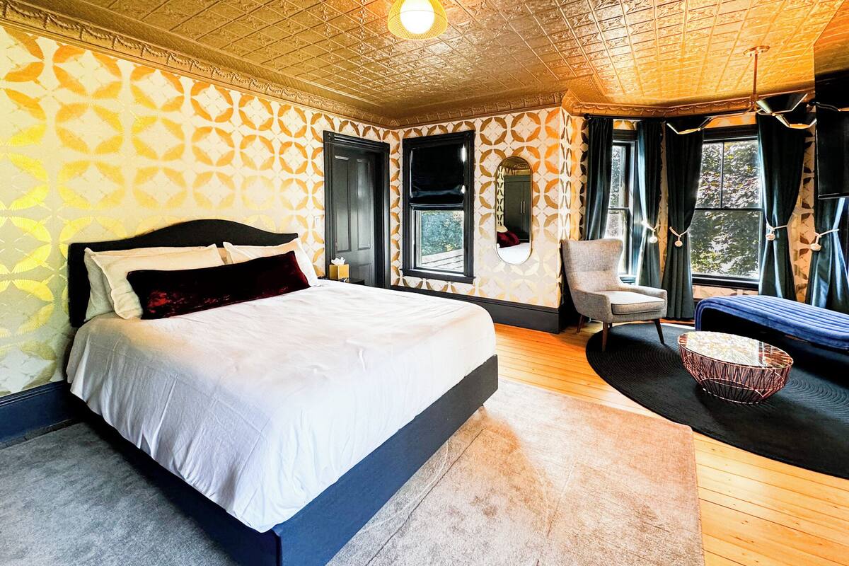 Une chambre à coucher avec un grand lit, une chaise, une petite table et une fenêtre avec des rideaux.