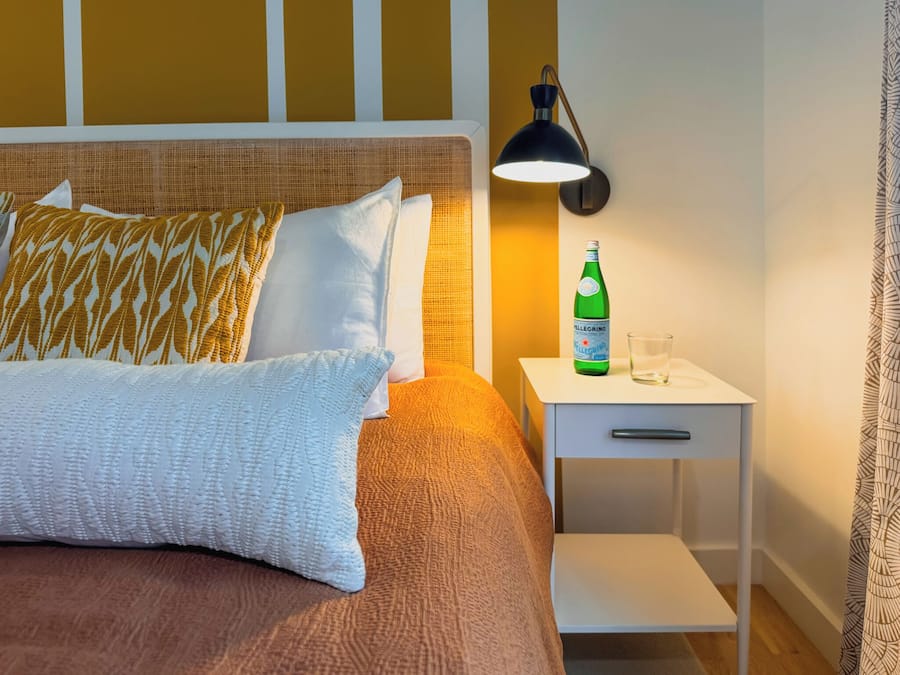 Una cama bien hecha con almohadones estampados, una mesita de noche con una botella y un vaso, y una lámpara de pared.