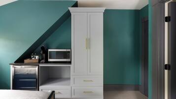 Ein Schlafzimmer mit türkisblauen Wänden, einem weißen Kleiderschrank mit goldenen Griffen, einem Bett mit weißer Bettwäsche und einer kleinen Küchenzeile mit einer Mikrowelle.