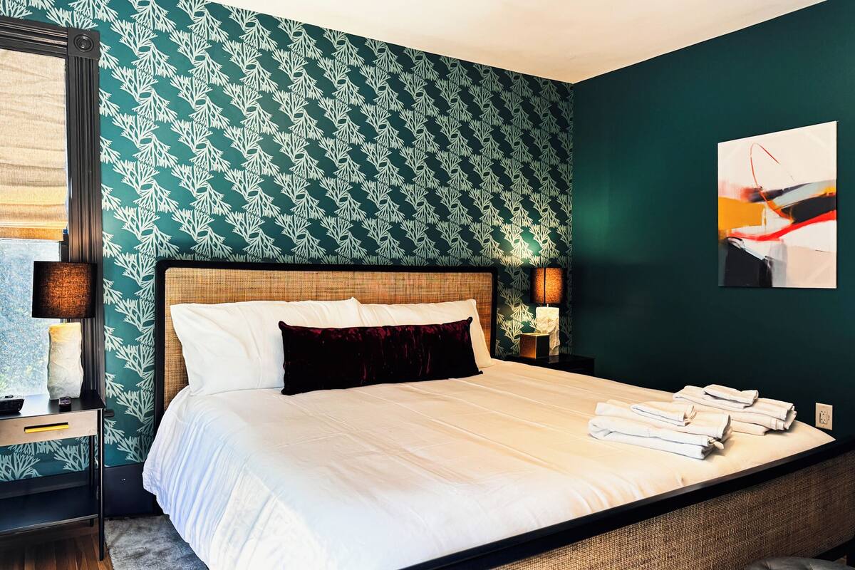 Un lit bien fait, avec du linge de lit blanc et un coussin rouge foncé, une table de chevet avec une lampe, et un mur orné d’un motif végétal.