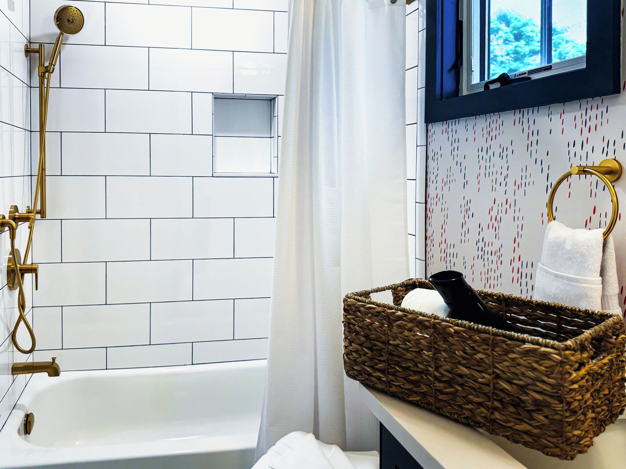 Un baño con pared de azulejos blancos, cabezal de ducha dorado, bañera, cesta de mimbre y toallero.