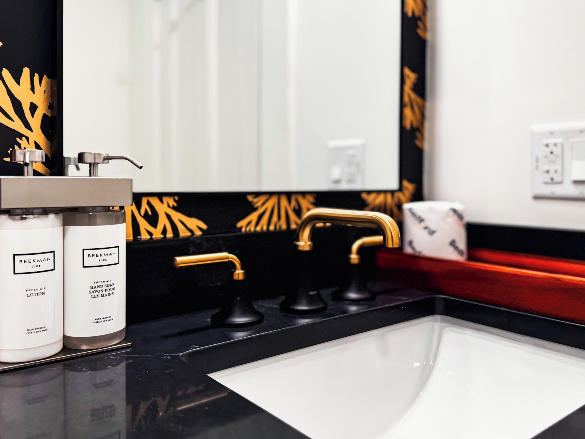 Une salle de bain avec un comptoir noir, un robinet doré et deux flacons de savon pour les mains de la marque Beekman.