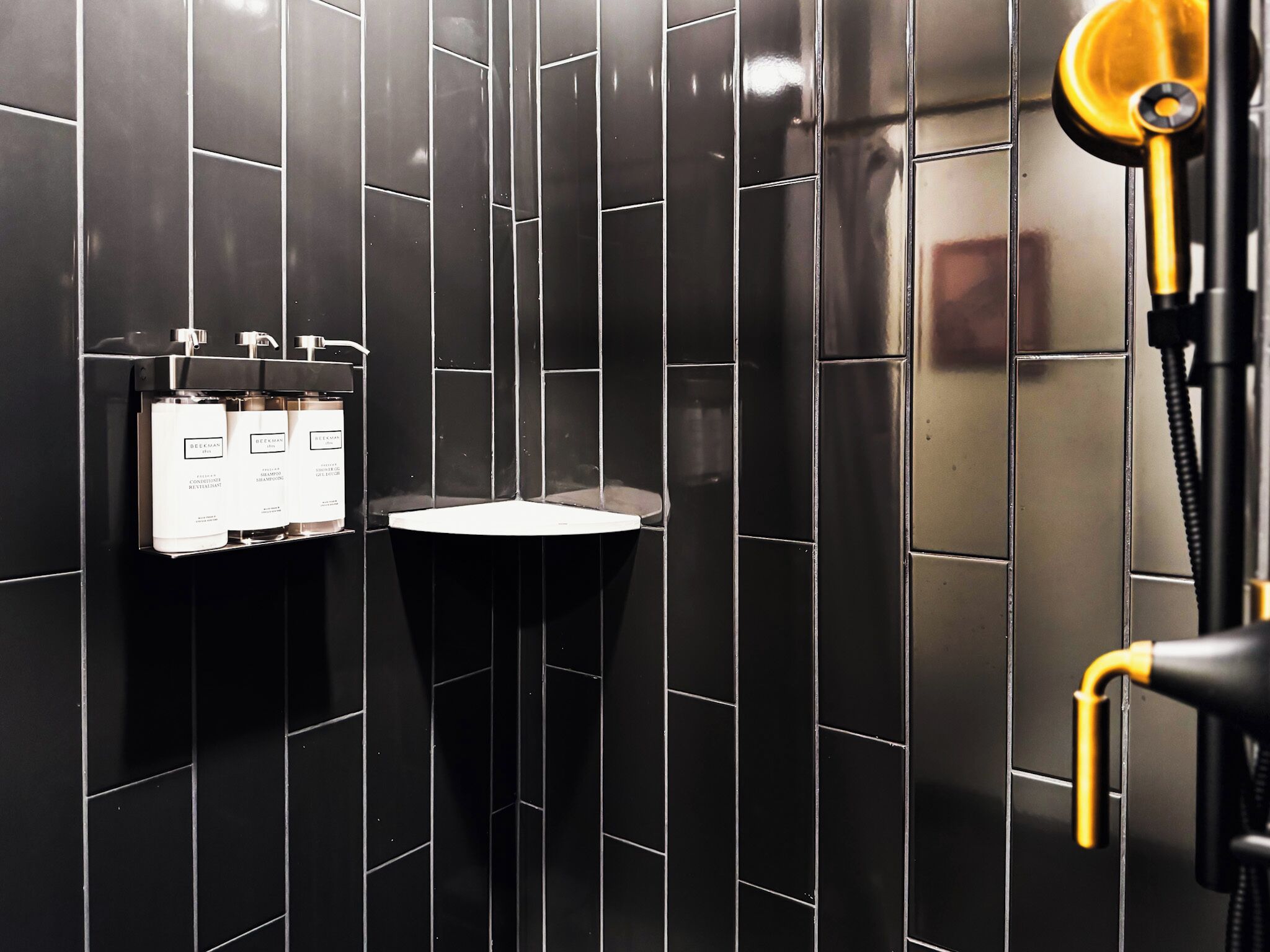 Un baño moderno con paredes de azulejos negros, un lavamanos blanco y un cabezal de ducha dorado.
