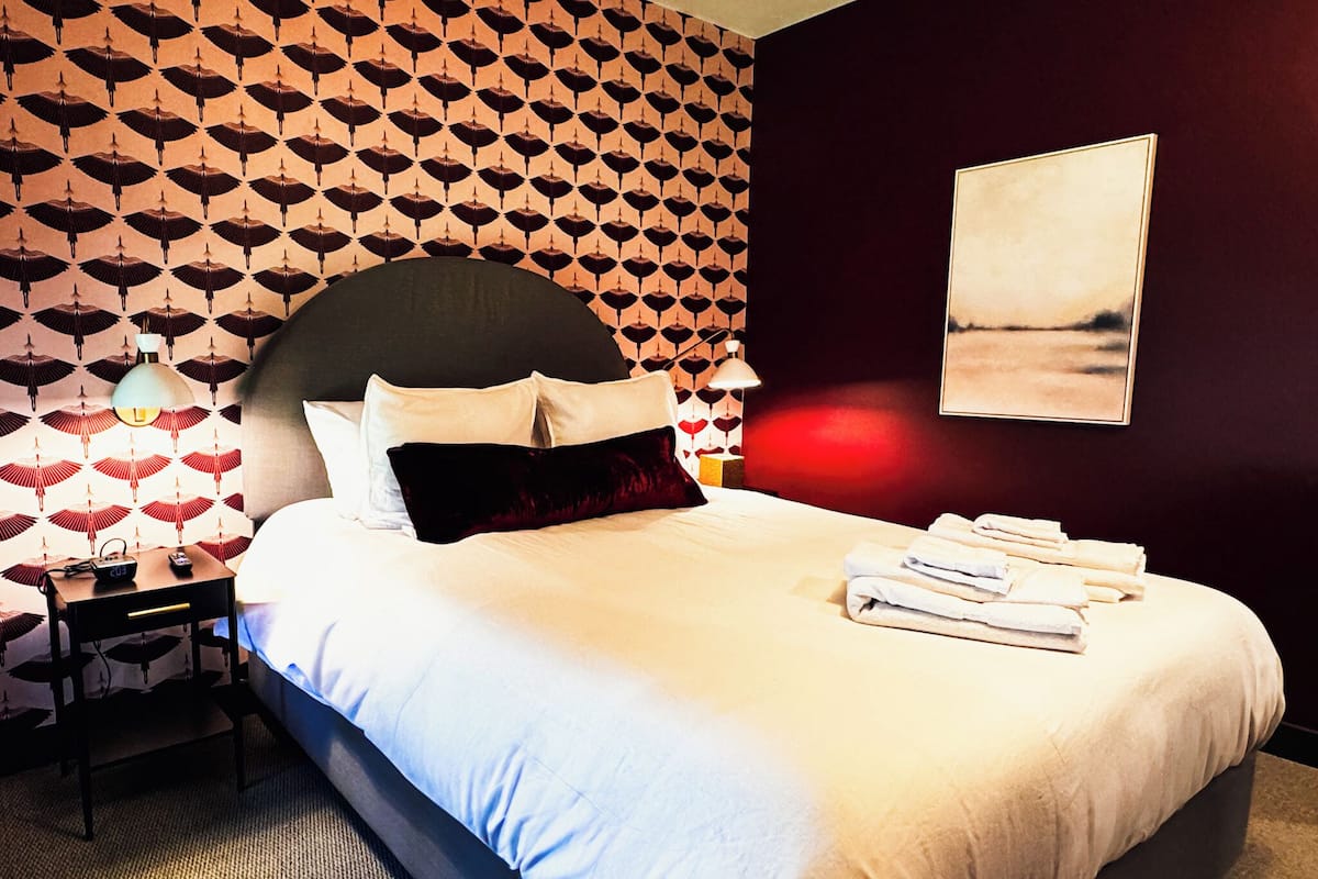Une chambre d’hôtel avec un grand lit, une table de chevet, une lampe et un mur recouvert d’un papier peint à motifs.