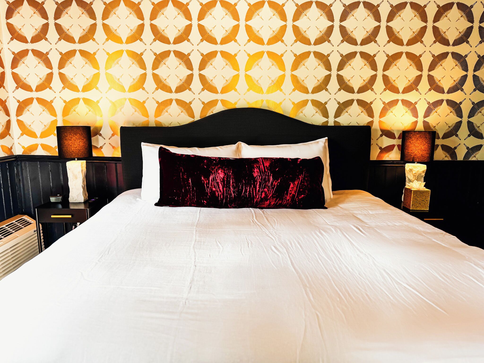 Ein ordentlich bezogenes Bett mit schwarzem Kopfteil, weißen Leinentüchern und einem roten Kissen mit Blumenmuster. Zwei Nachttischlampen mit braunen Lampenschirmen und eine kleine Statue stehen auf dem Nachttisch. Die Wand ist mit geometrischem Wandpapier gestaltet.
