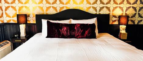 Cama bem arrumada com cabeceira preta, roupa de cama branca e almofada floral vermelha. Duas luminárias de cabeceira com abajures marrons e uma pequena estátua no criado-mudo. O papel de parede da parede apresenta um padrão geométrico.