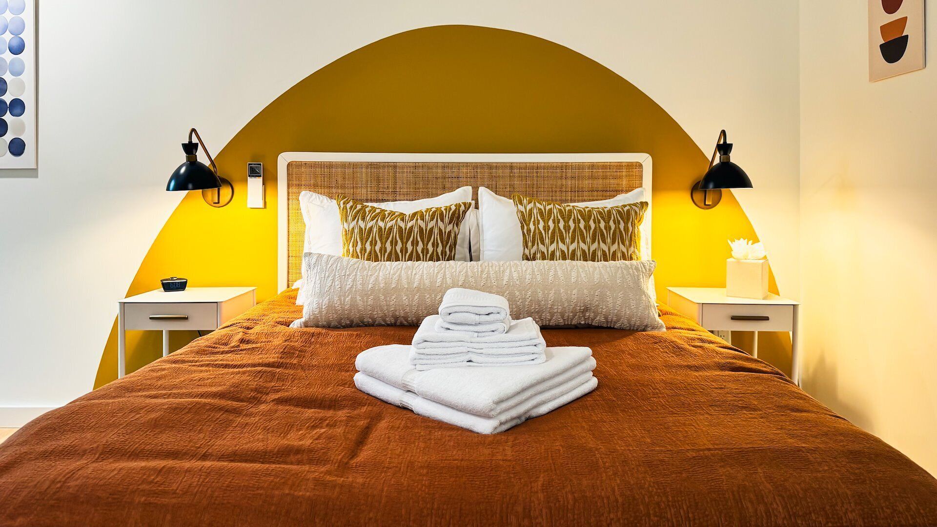 Una cama con sábanas bien ordenadas, una manta marrón, almohadas blancas y una pila de toallas blancas. Hay dos mesitas de noche, cada una con una lámpara. La habitación tiene una pared de acento amarilla y un arco grande y curvo sobre la cama.