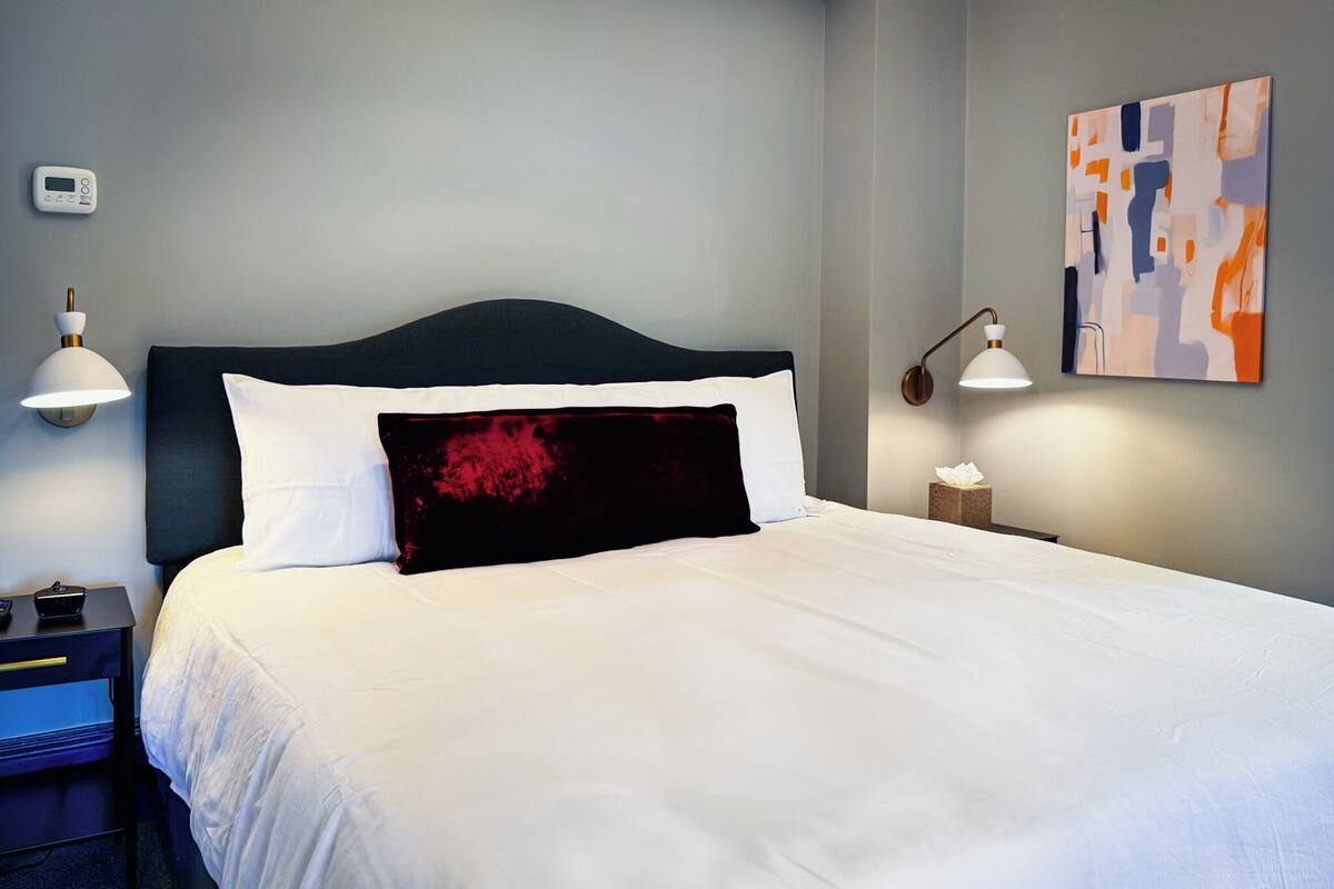 Un lit bien fait, avec du linge de lit blanc et un coussin décoratif noir et rouge, une table de chevet avec une lampe, et un tableau abstrait accroché au mur.