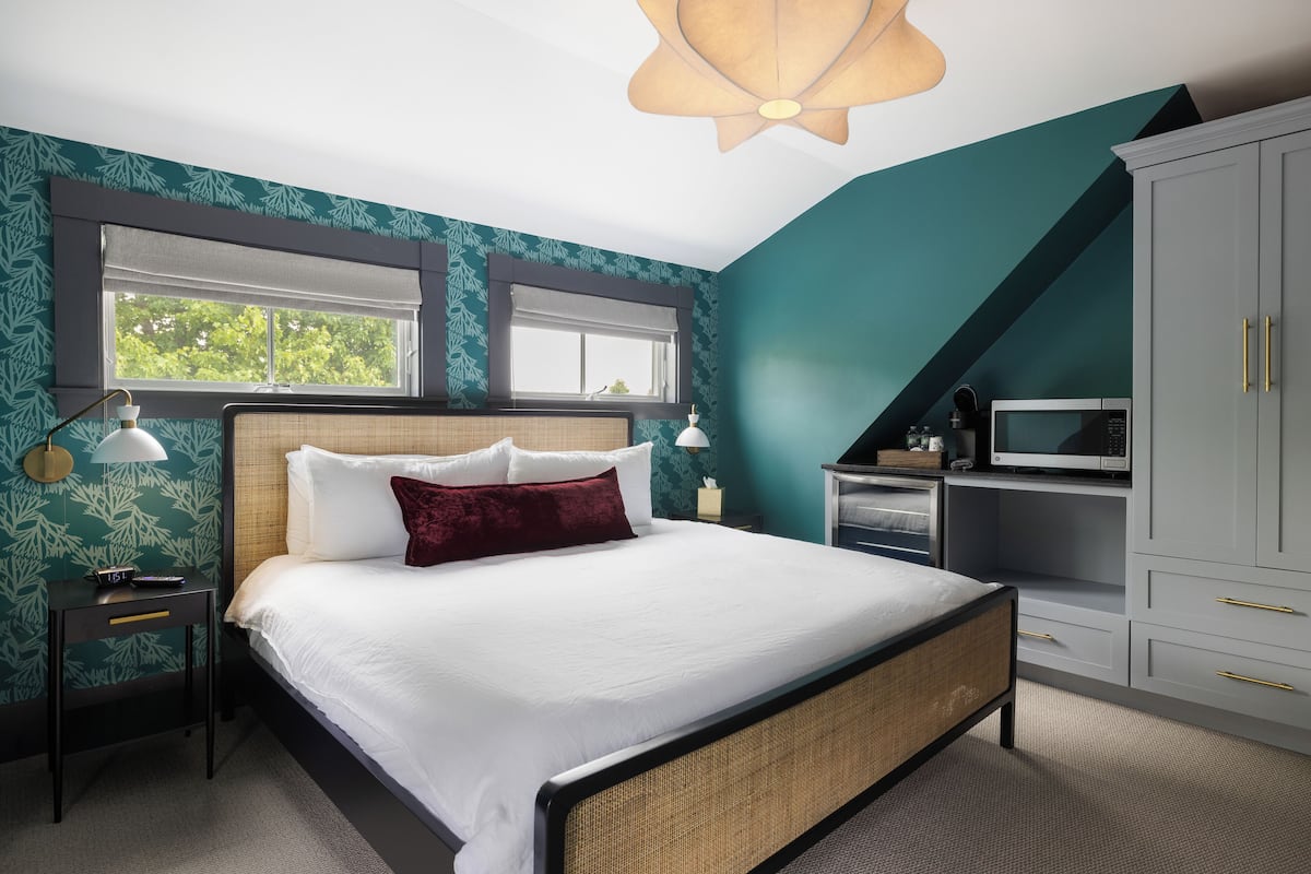 Une chambre à coucher avec un grand lit, une table de chevet, une lampe, une armoire et une fenêtre donnant sur des arbres.