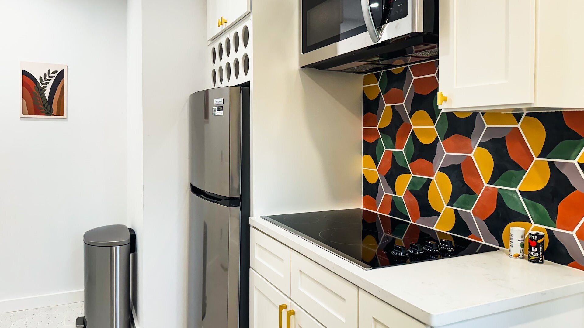 Cozinha com geladeira de aço inoxidável, micro-ondas, fogão e painel de azulejos hexagonais coloridos.