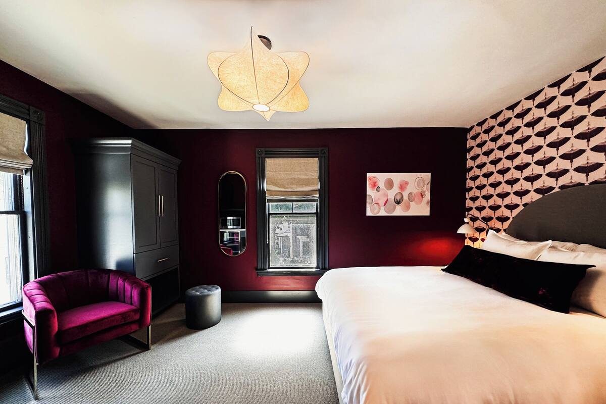 Une chambre à coucher moderne avec un grand lit, un fauteuil rouge, une armoire et un luminaire au plafond original.