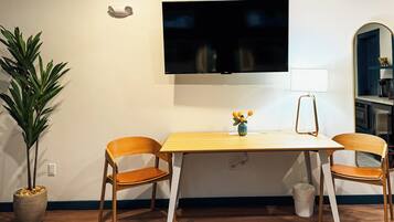 Un salon moderne avec un téléviseur à écran plat fixé au mur, deux fauteuils en bois, une petite table sur laquelle est posé un vase de fleurs, un lampadaire et une plante en pot.