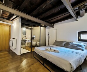Une chambre avec un lit, un plancher en bois, un plafond avec des poutres apparentes, une porte-fenêtre coulissante donnant sur une salle de bain et une fenêtre avec vue.