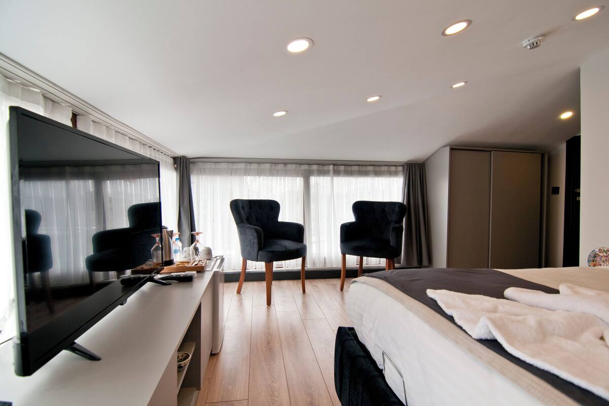 A modern hotel room with a flat-screen TV, a desk with a computer, two black armchairs, a bed with a white comforter, and a large wardrobe.