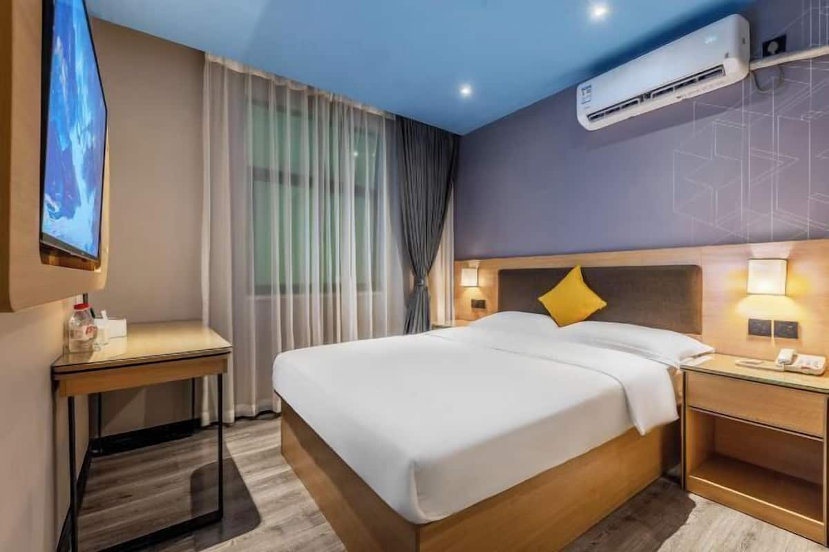 A hotel room with a bed, bedside table, lamp, air conditioning unit, and a TV.