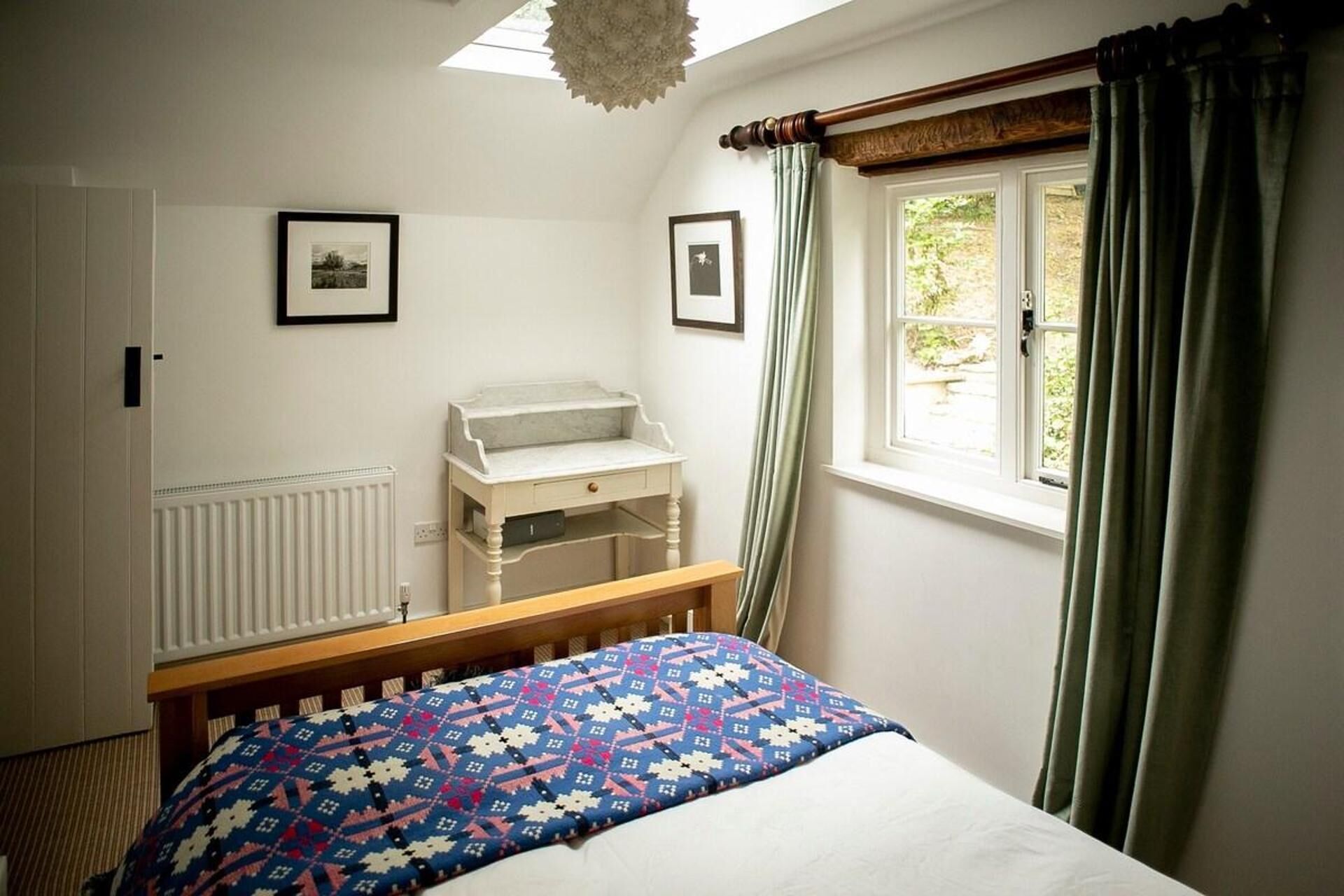 A bedroom with a bed, a dresser, a radiator, a window with curtains, and two framed pictures on the wall.