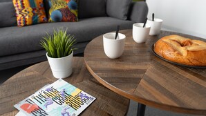 A cozy living room with a grey sofa, colorful pillows, a wooden coffee table with a magazine, a potted plant, and a braided loaf of bread.