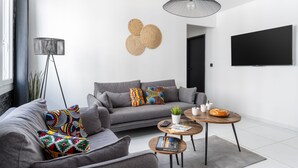 A modern living room with a grey sofa, colorful cushions, a round coffee table, a floor lamp, and a flat-screen TV mounted on the wall.