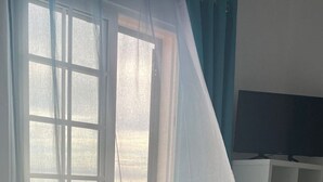 A room with a window covered by sheer curtains, a television on a stand, and a white shelving unit.