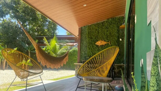 Un espace extérieur abrité comprenant deux fauteuils en osier jaune, un hamac suspendu et un mur vert orné d’une décoration représentant un poisson.