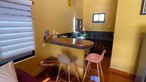 Une cuisine compacte avec un coin bar, deux tabourets et une fenêtre équipée de stores.