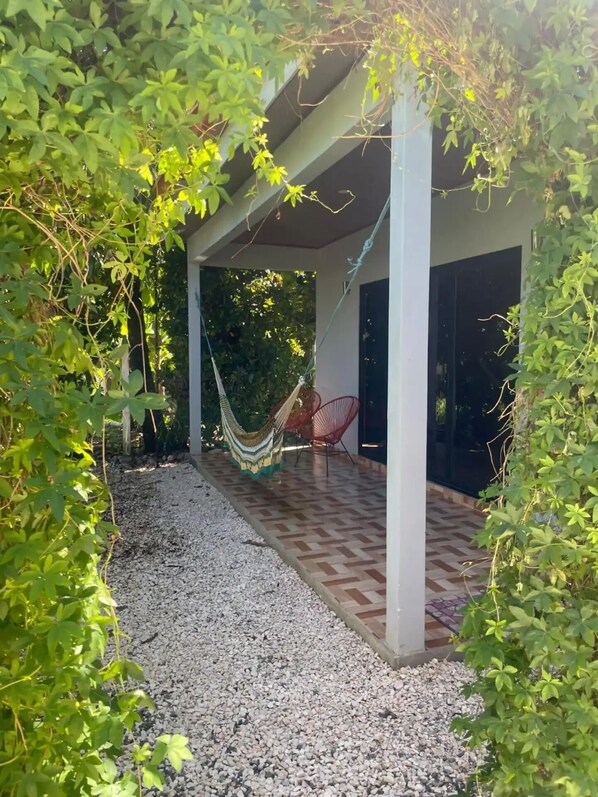 A covered outdoor area with a hammock, a red chair, and a paved floor.