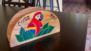 Une table en bois ornée d’un perroquet aux couleurs vives et sur laquelle est posée une plaque nominative. En arrière-plan, on aperçoit un coin cuisine avec un comptoir, des chaises et une fenêtre donnant sur un espace verdoyant.