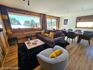 A modern living room with a sofa, coffee table, and armchair, a dining area with a table and chairs, and large windows offering a view of trees and a building. - Appartement  A-04  T4 (Font-Romeu-Odeillo-Via)
