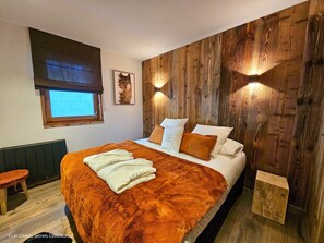 A bedroom with a wooden wall panel, a bed with white and brown bedding, a window with a view, and a small stool.