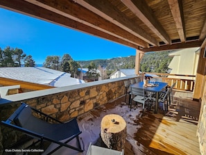 Una terraza de madera con un muro de piedra, una mesa con sillas y vistas a tejados y árboles cubiertos de nieve.
