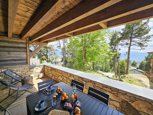 An outdoor dining area with stone walls and wooden beams, overlooking a scenic landscape with trees and mountains.