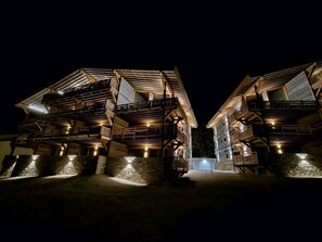 A night view of a traditional wooden chalet with multiple balconies and stone foundations. - Appartement B-03 T2 Cabine (Font-Romeu-Odeillo-Via)