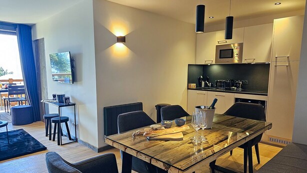 A modern dining area with a wooden table, black chairs, and a kitchen in the background. - Appartement B-12 T2 Cabine (Font-Romeu-Odeillo-Via)