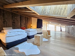 Habitación con techos altos y vigas de madera, dos camas con ropa blanca, un taburete de madera pequeño y una alfombra de piel en el suelo.