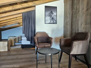 A loft-style room with a sloped ceiling, wooden beams, a large window with curtains, two leather armchairs, a small round table, and a framed mountain photograph on the wall.