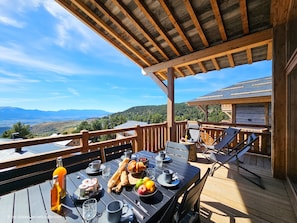 Una terraza de madera con una mesa dispuesta para el desayuno, con vistas a un paisaje montañoso.