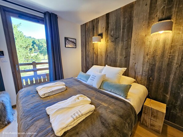 A bedroom with a large bed, wooden headboard, two bedside lamps, and a view of a wooden deck and trees.